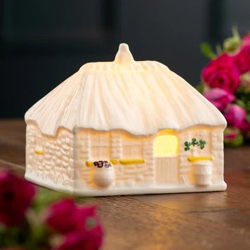 Cottage-shaped night light with a warm glow on a wooden surface with flowers.