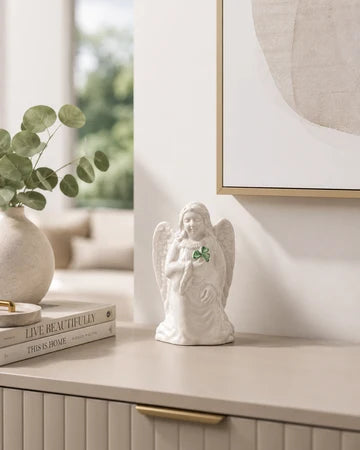 A small white angel figurine with wings and a green shamrock accent stands on a light beige console table in a styled interior setting.