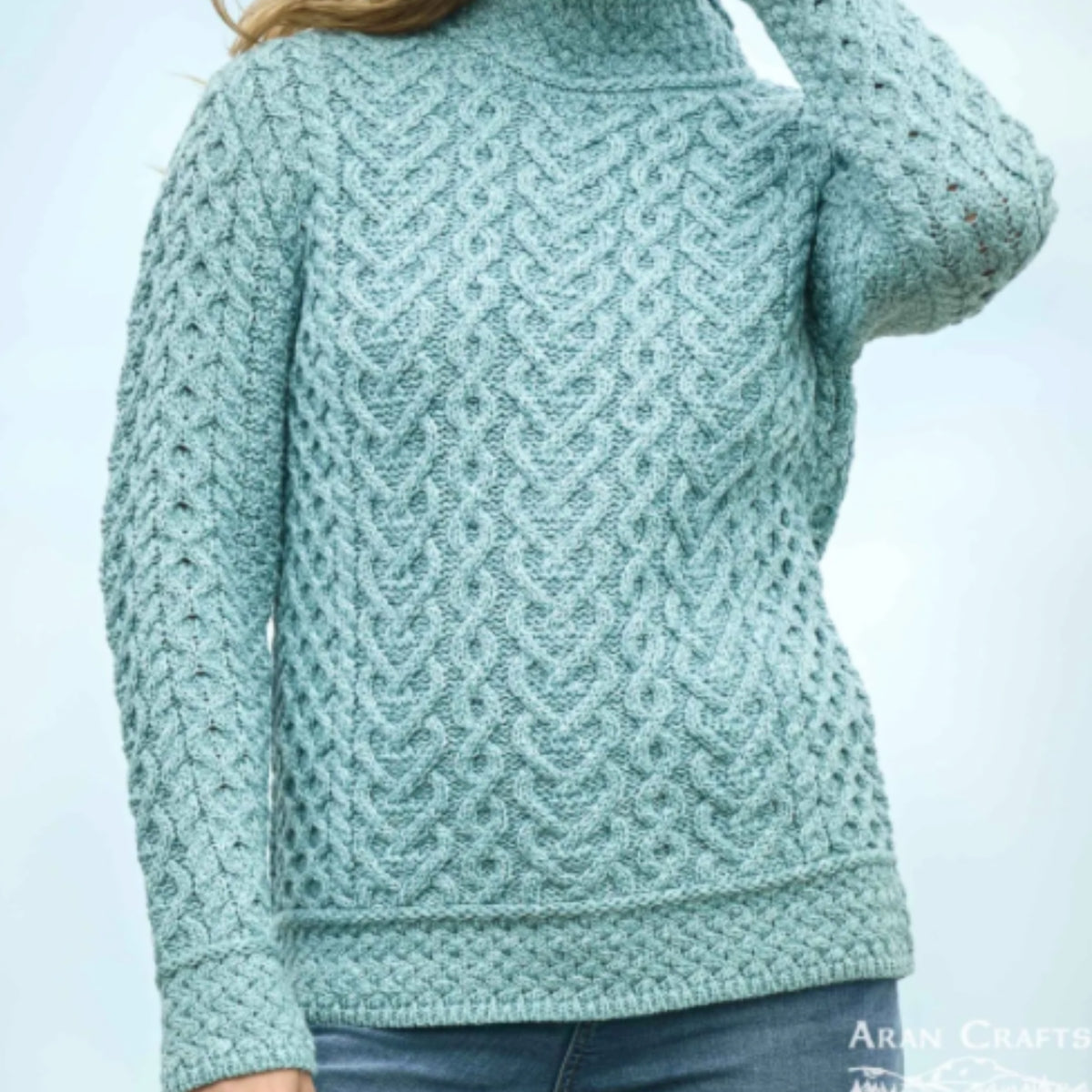 A woman wearing a light blue-green cable-knit turtleneck sweater with a textured openwork pattern, paired with blue jeans. The brand name 'Aran Crafts' is visible in the bottom right corner.