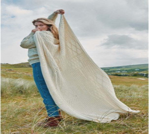 Woman holding a large white knitted blanket in a grassy field with a scenic background