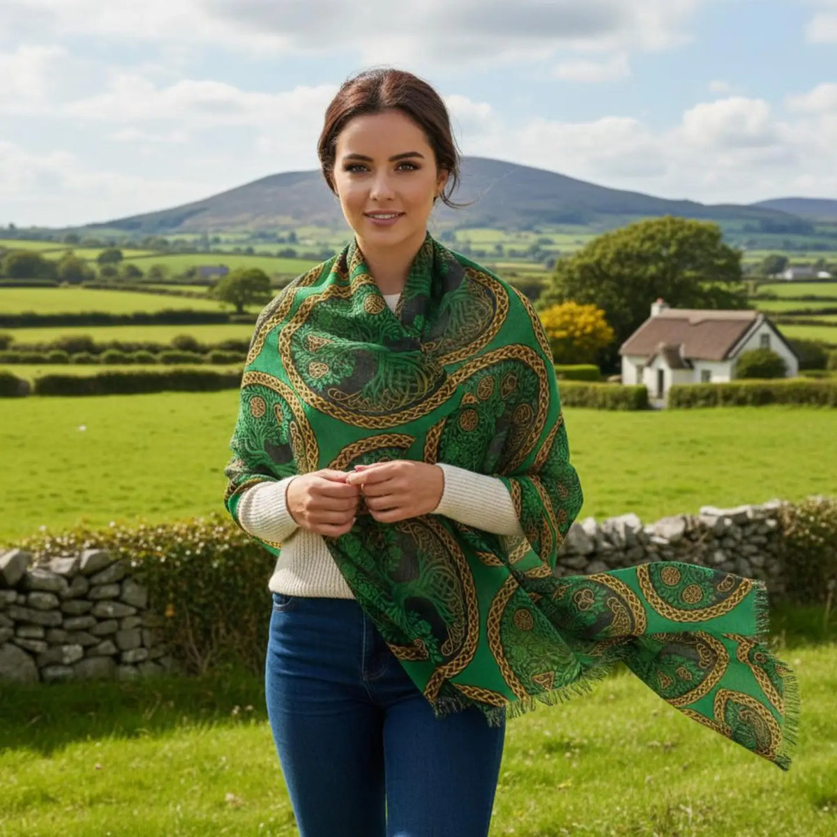 A woman standing outdoors in a green rural landscape, wearing a green shawl with a gold and dark paisley pattern over a white sweater and blue jeans.