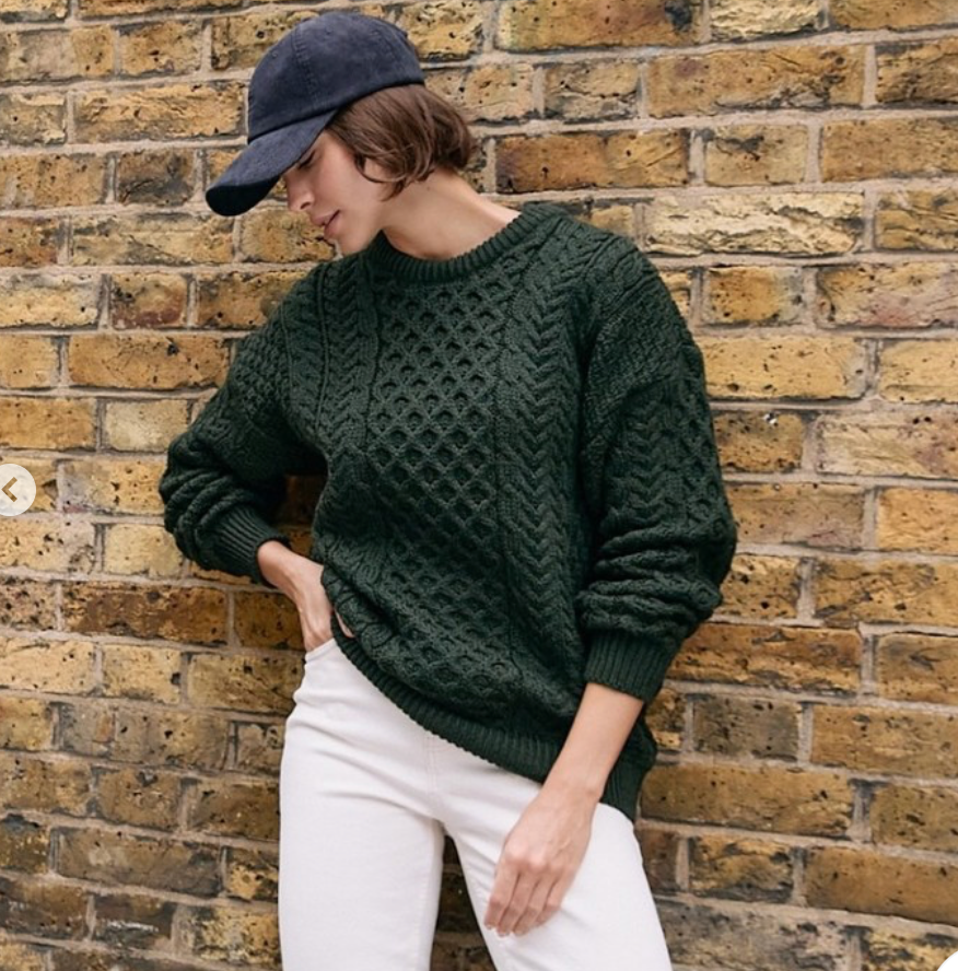 A woman wearing a dark forest green cable-knit sweater and white pants, leaning against a yellow brick wall and wearing a navy baseball cap.