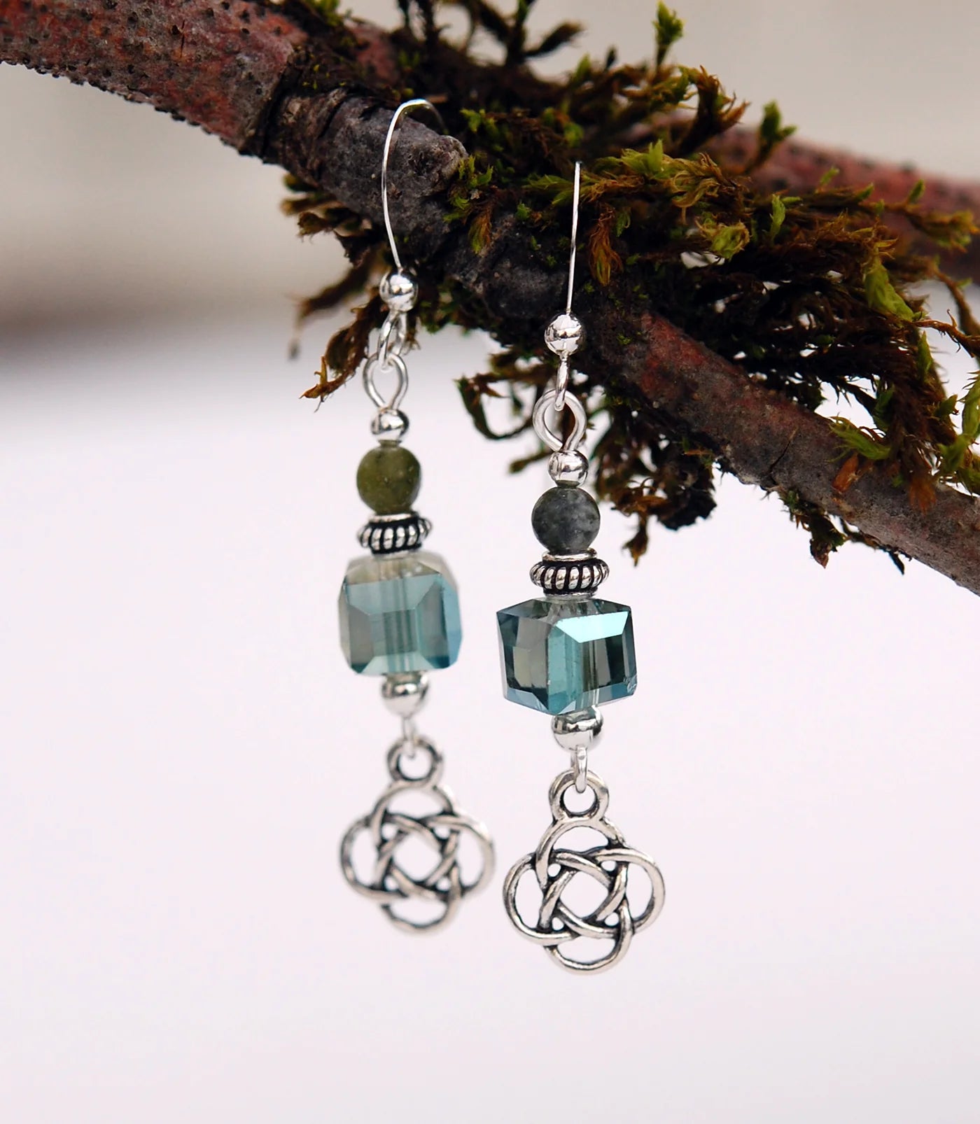 Pair of earrings with green and blue beads and silver Celtic knot designs on a branch background.