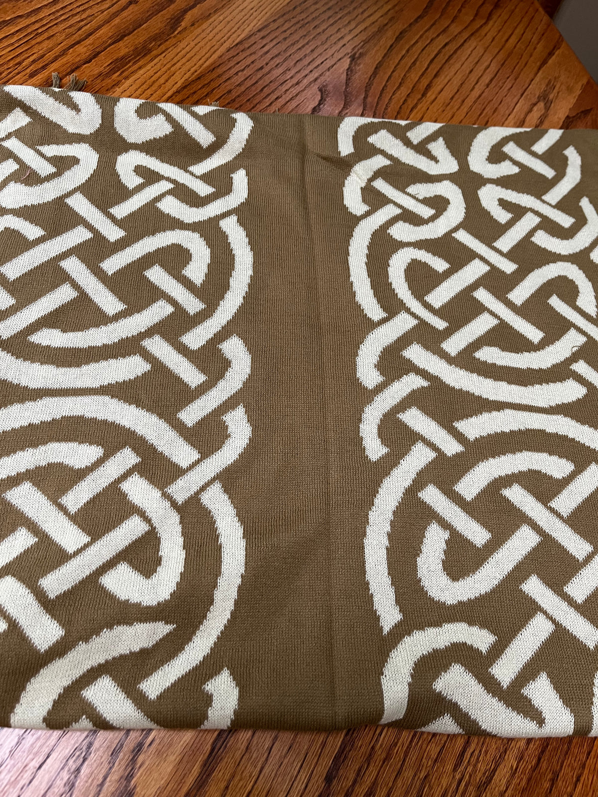 A close-up of a woven textile with a repeating interlocking knot pattern in white over a medium brown background, laid flat on a wooden surface.