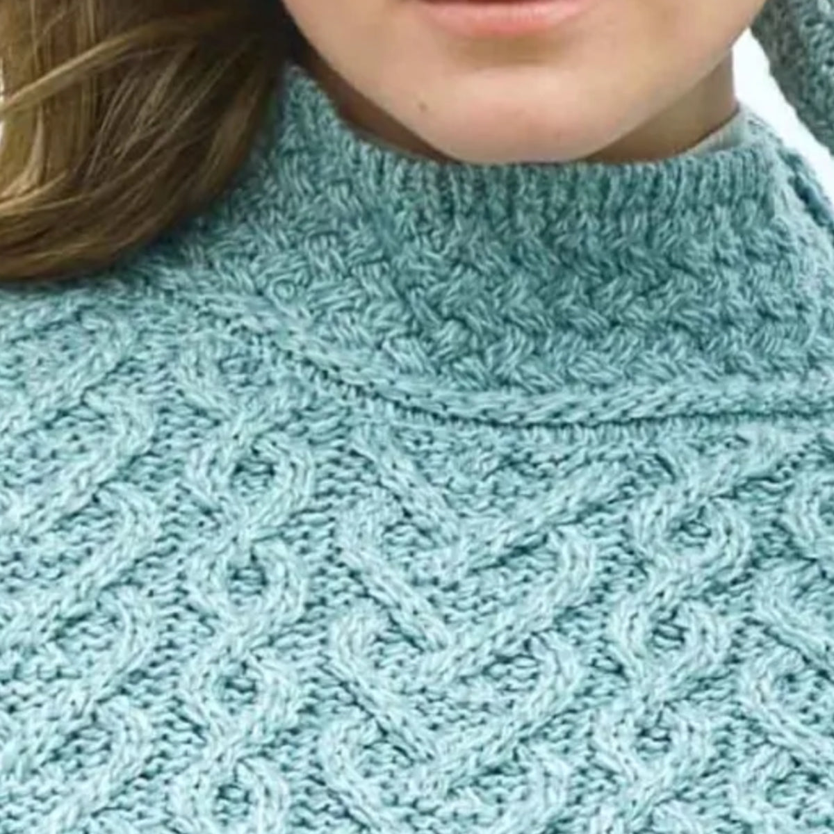 Close-up of a person wearing a chunky knit turtleneck sweater in a dusty light blue-green color, showing the cable-knit texture and the model's chin and hair.