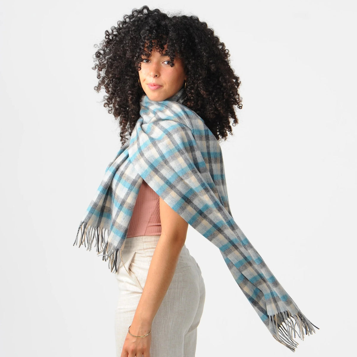 A woman with curly black hair models a light blue, gray, and white plaid fringed scarf wrapped around her shoulders, paired with a pink top and white pants against a plain white background.
