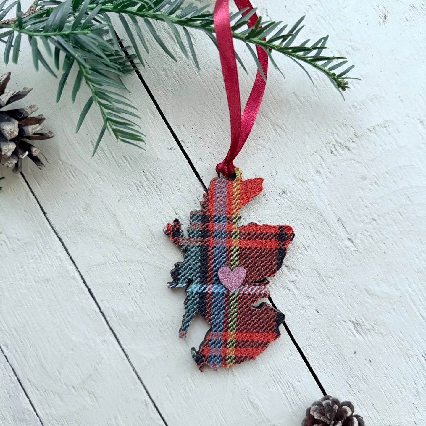 A Scotland map-shaped fabric ornament in a red tartan plaid pattern with a small pink heart in the center, hanging from a red ribbon on a white wooden surface with pine branches and pinecones.