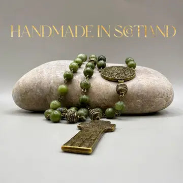 A handmade green stone rosary with bronze-toned metal beads, a round medallion, and a cross pendant, arranged on a smooth light stone against a neutral gray background. Gold text at the top reads 'HANDMADE IN SCOTLAND'.