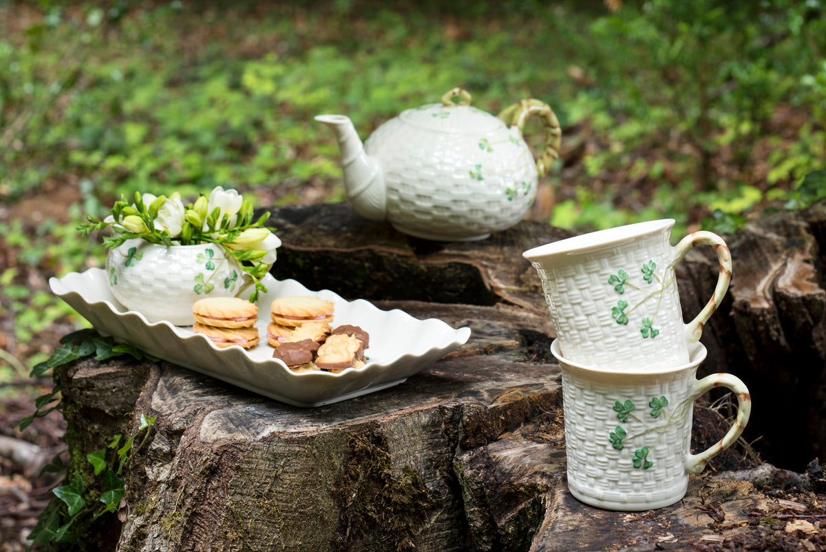 A set of white ceramic tableware with green clover motifs, including a teapot, stacked mugs, and a serving dish with cookies and flowers, arranged on a tree stump in a forest setting.