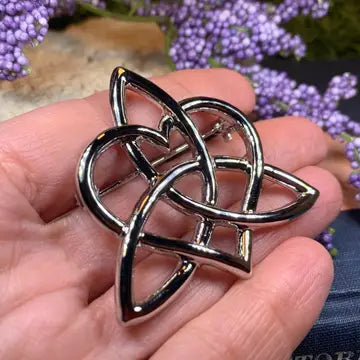 A polished pewter Celtic triquetra knot brooch held in a person's hand, with blurred purple flowers in the background.