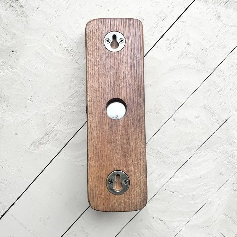 A rectangular wooden doorbell push button with a warm brown wood grain finish, featuring a central circular cutout and two silver metal keyhole-shaped screws at the top and bottom, shown on a white wood plank background.