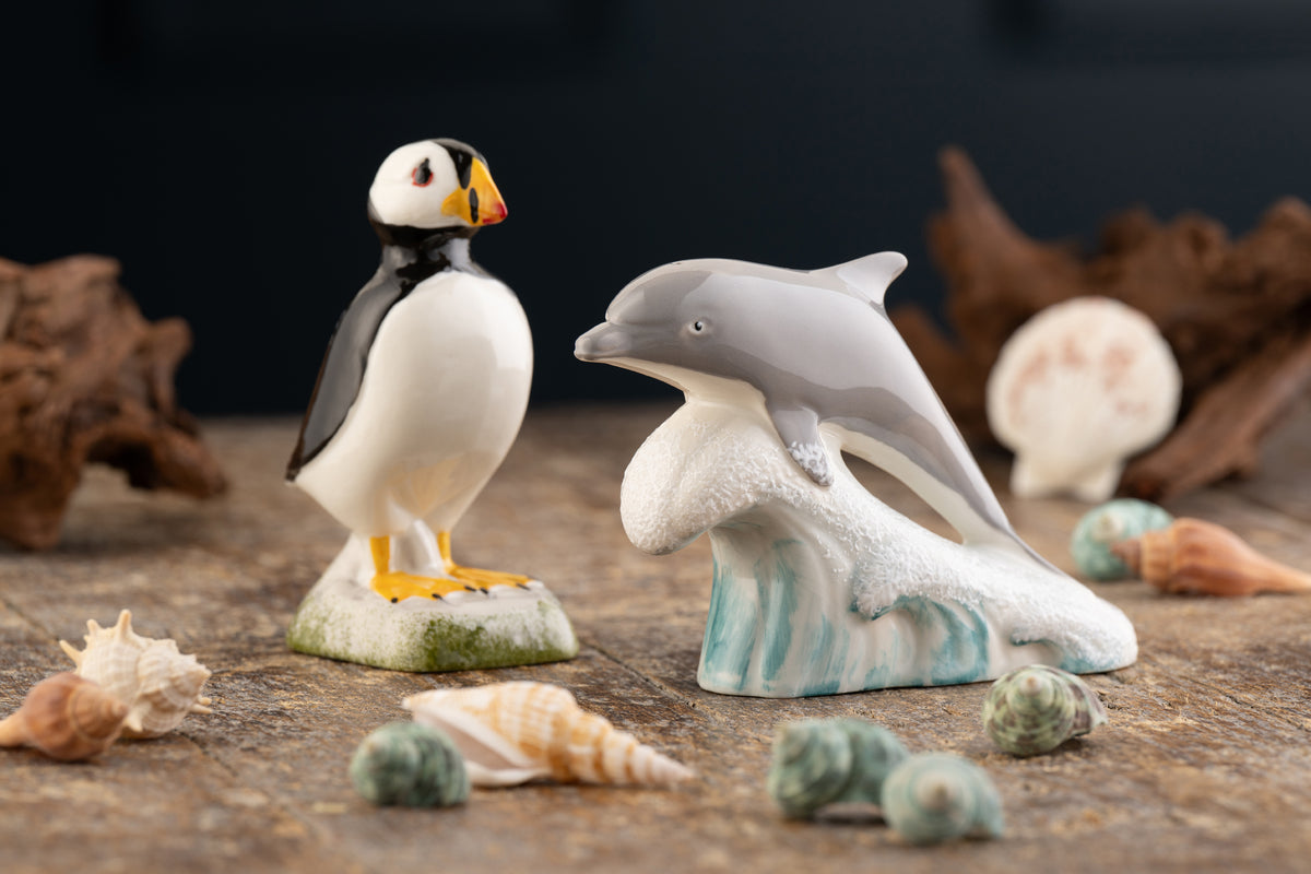 A ceramic figurine set featuring a puffin standing on a rock and a dolphin emerging from waves, displayed on a rustic wooden surface with seashells and driftwood.
