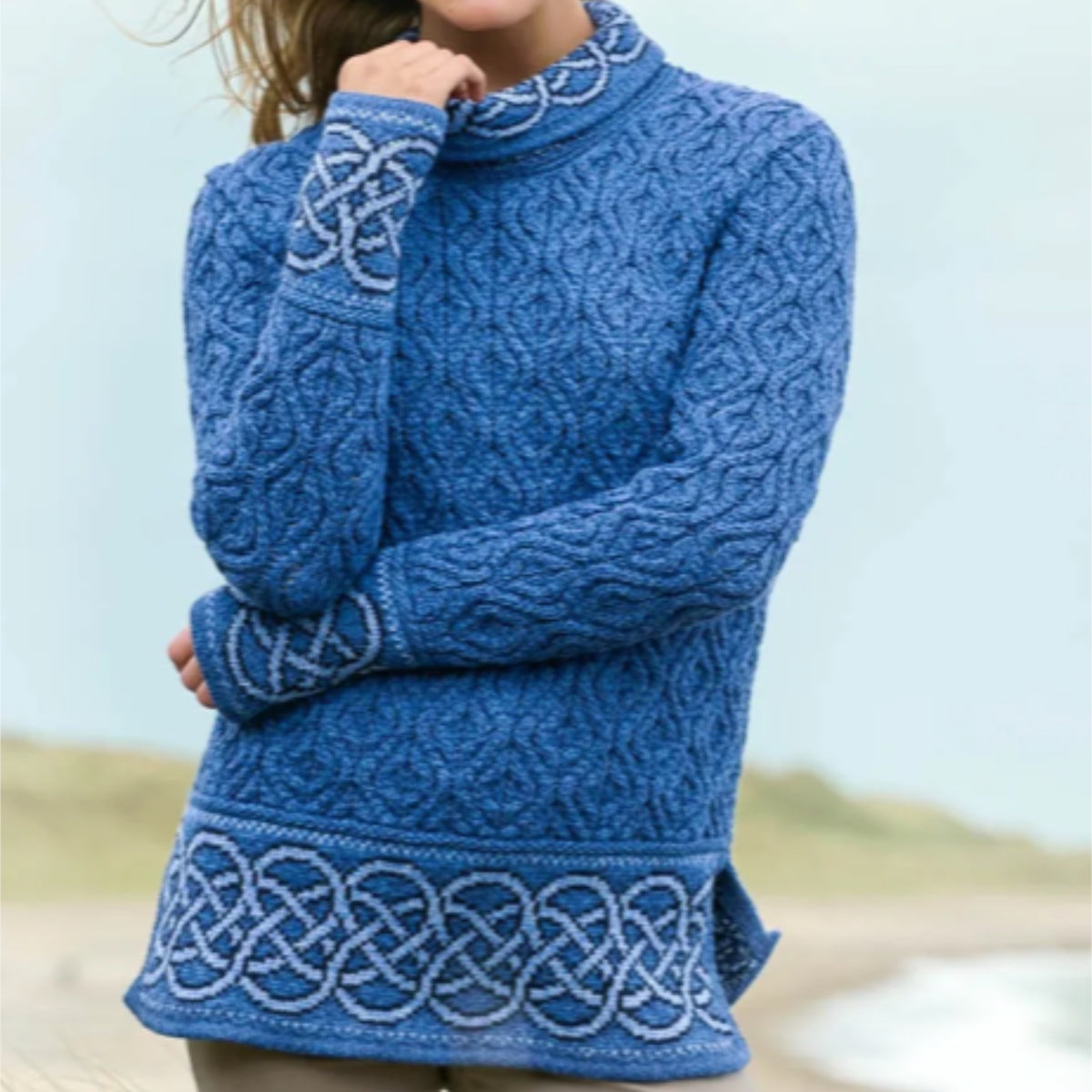 A woman wearing a blue cable-knit mock-neck sweater with white Celtic knot patterns on the cuffs and hem, photographed outdoors against a soft beach background.