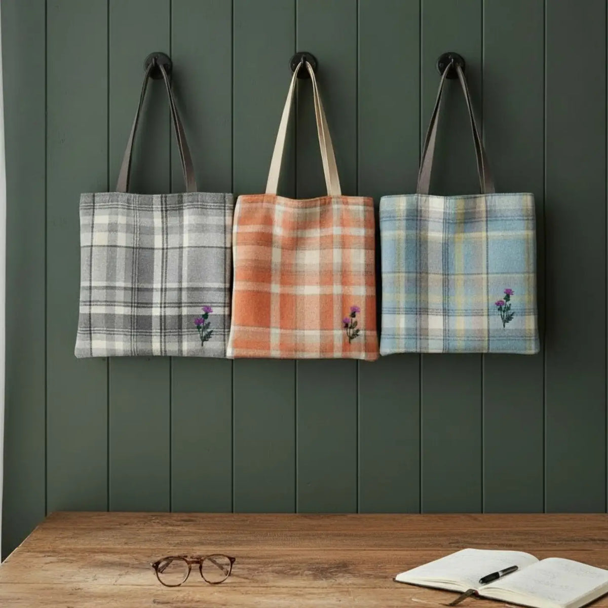 Three plaid fabric tote bags hang on a dark green paneled wall above a wooden table with an open notebook and glasses. The tote bags have dark brown or beige handles and small embroidered purple flowers near the bottom left corner.