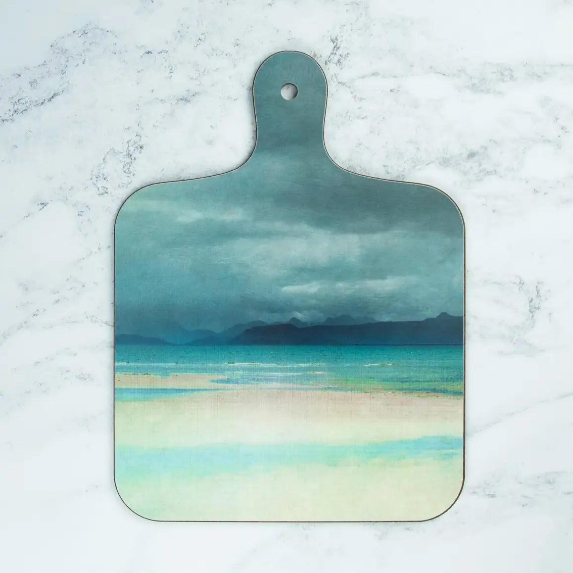 A wooden cutting board with a handle, featuring a painted beach and ocean scene in shades of teal, blue, and pale yellow, displayed on a white marble surface.