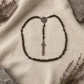 A dark brown beaded rosary necklace with a silver-toned crucifix pendant and a round medallion, arranged on a textured cream linen fabric with decorative stones and dried flowers.