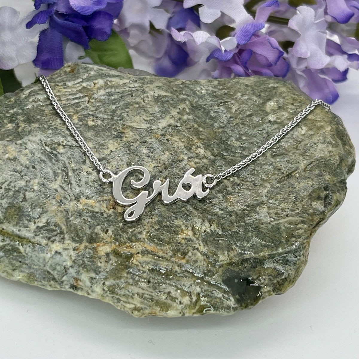 A silver script name necklace reading 'Grá' is draped over a textured gray stone, with purple and white flowers blurred in the background.