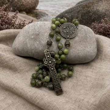 An olive green beaded rosary with an ornate bronze cross and medallion, draped over a smooth gray stone on a beige linen cloth with rocks in the background.
