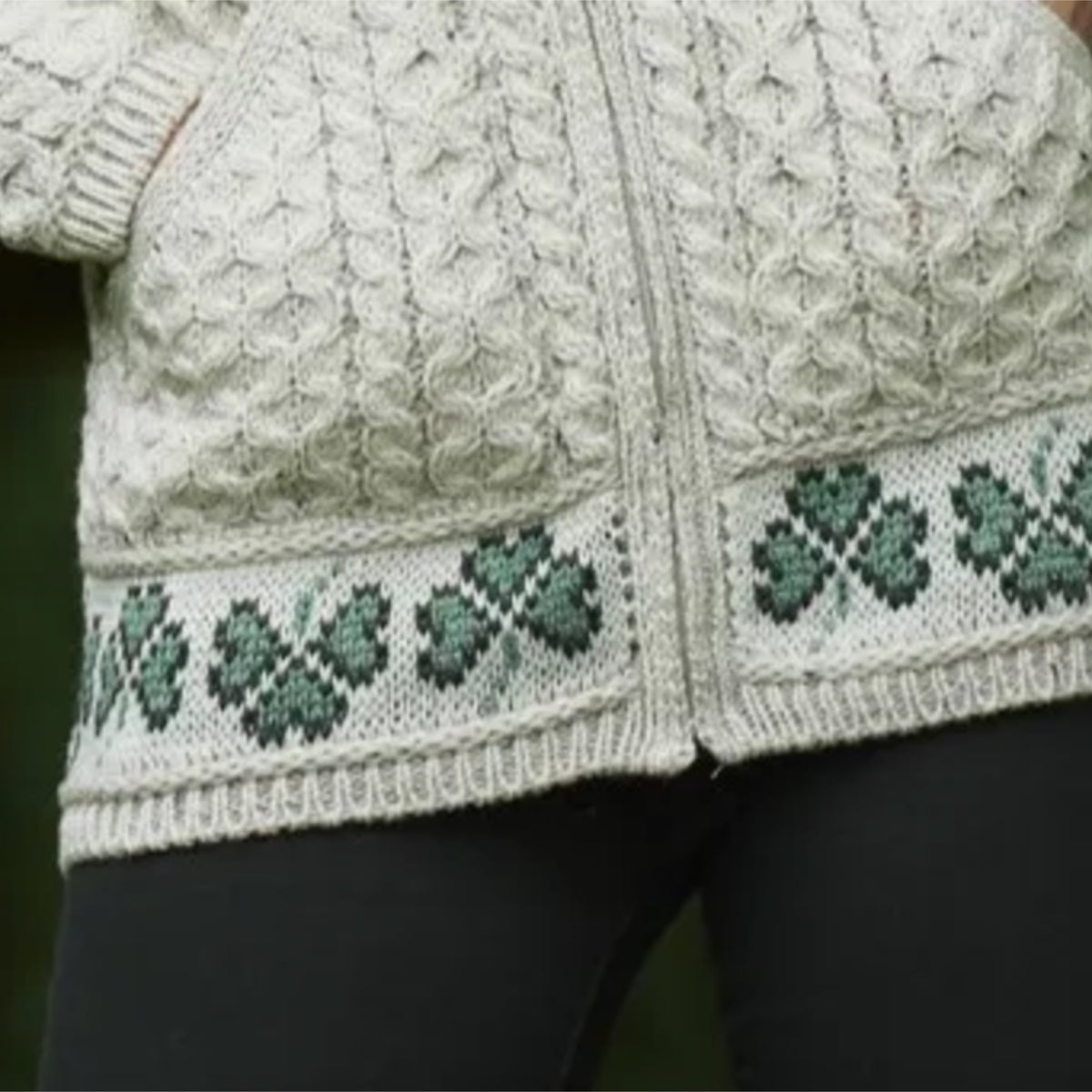 Close-up of a cream cable-knit zip-front cardigan with green shamrock patterns along the hem, worn over black leggings.