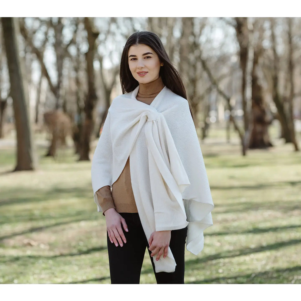 A woman wearing a cream-colored knit wrap or poncho over a tan turtleneck and black pants, standing in a park with blurred trees in the background.