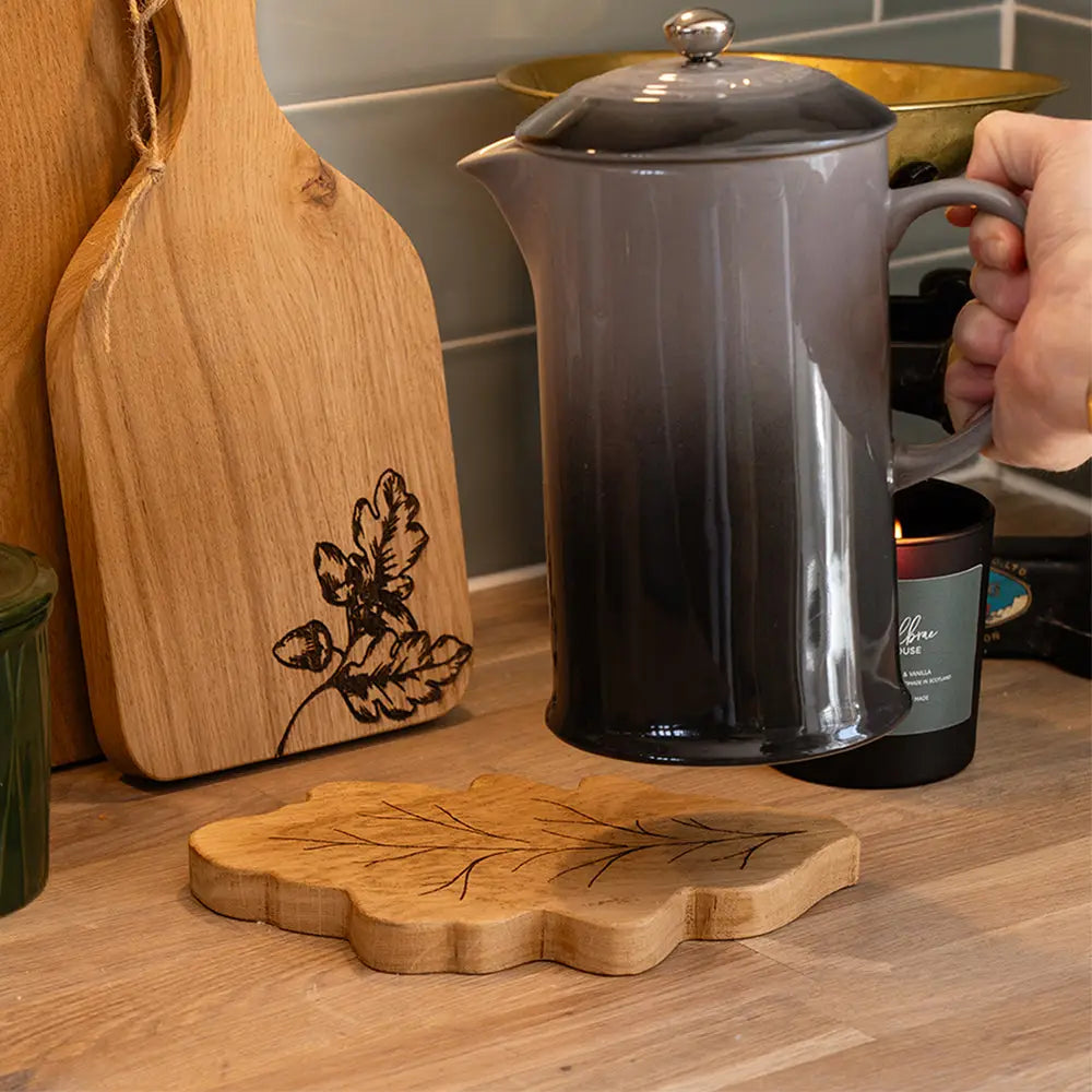 A dark gray to black gradient enamel coffee pot with a silver lid and handle, held by a hand on a wooden kitchen countertop. In the foreground, a leaf-shaped wooden trivet sits on the counter, with a wooden cutting board and other kitchen items in the background.