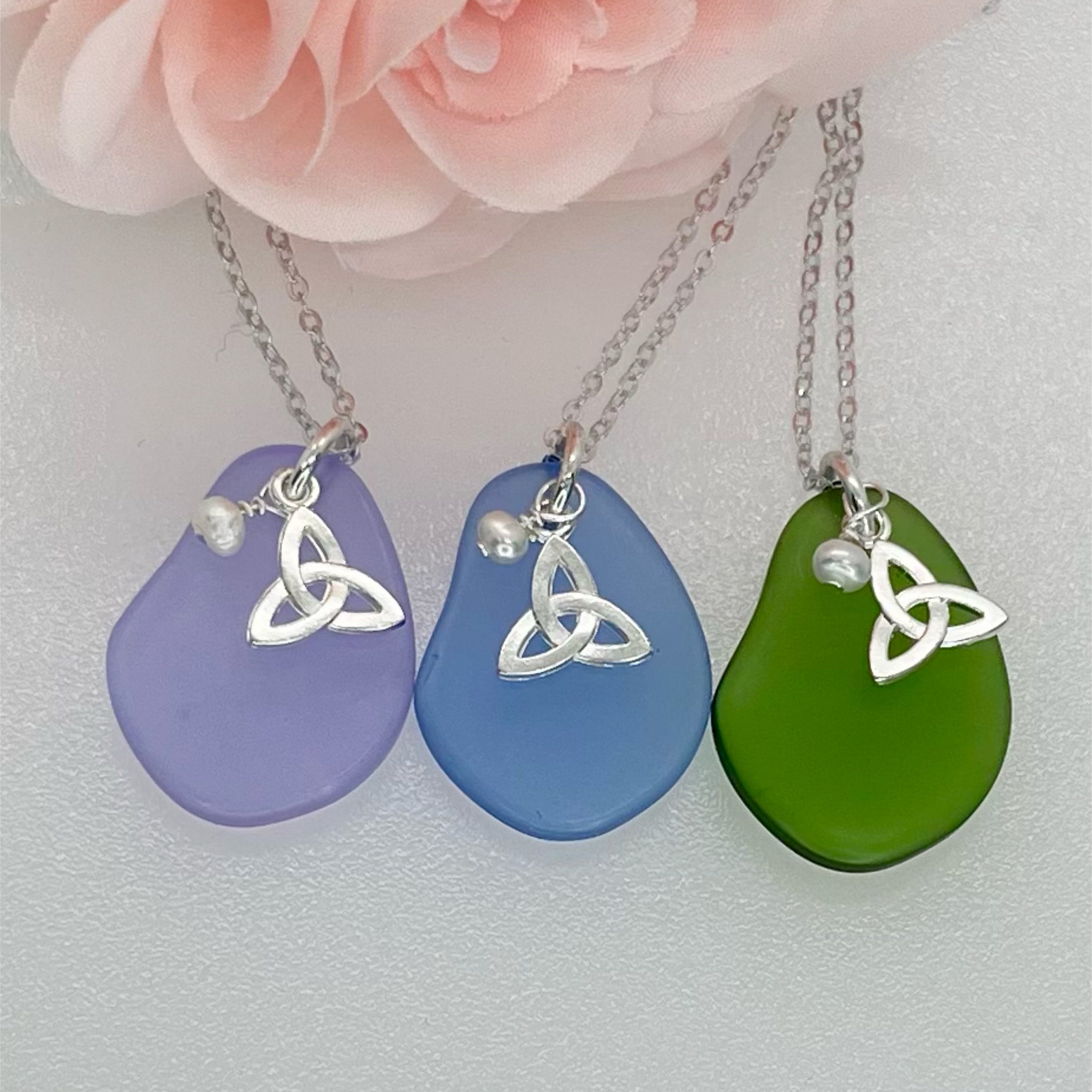 Three teardrop-shaped glass pendants in purple, blue, and green with silver Celtic knot designs on a light pink floral background.