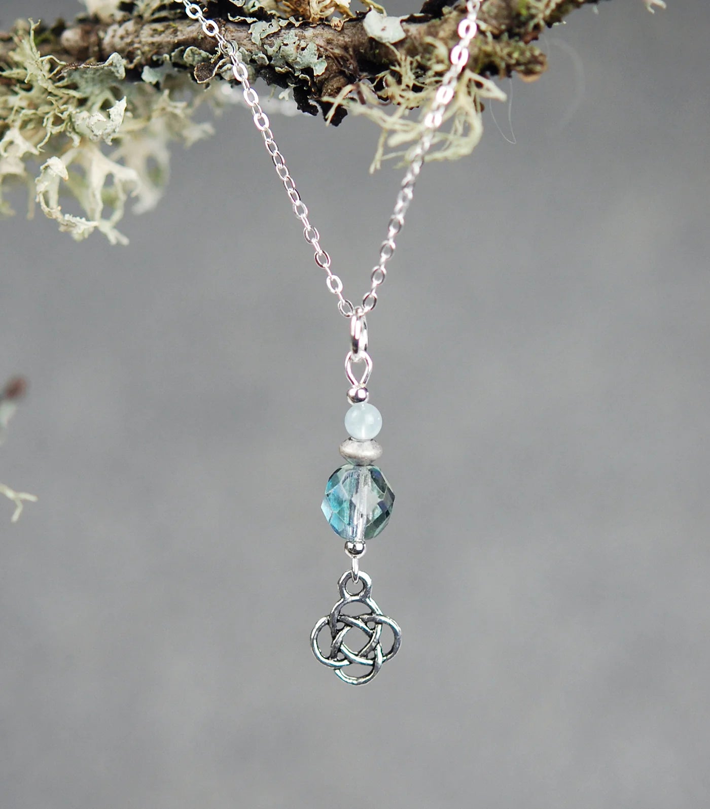 Necklace with a pendant featuring a Celtic knot design and a blue gemstone, hanging from a branch with lichen.