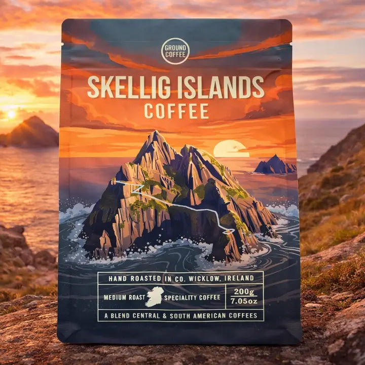 A stand-up coffee bag labeled Ground Coffee Skellig Islands Coffee, featuring an illustrated rocky island seascape at sunset. The package is photographed outdoors against a real coastal landscape with ocean, cliffs, and a glowing orange sky.