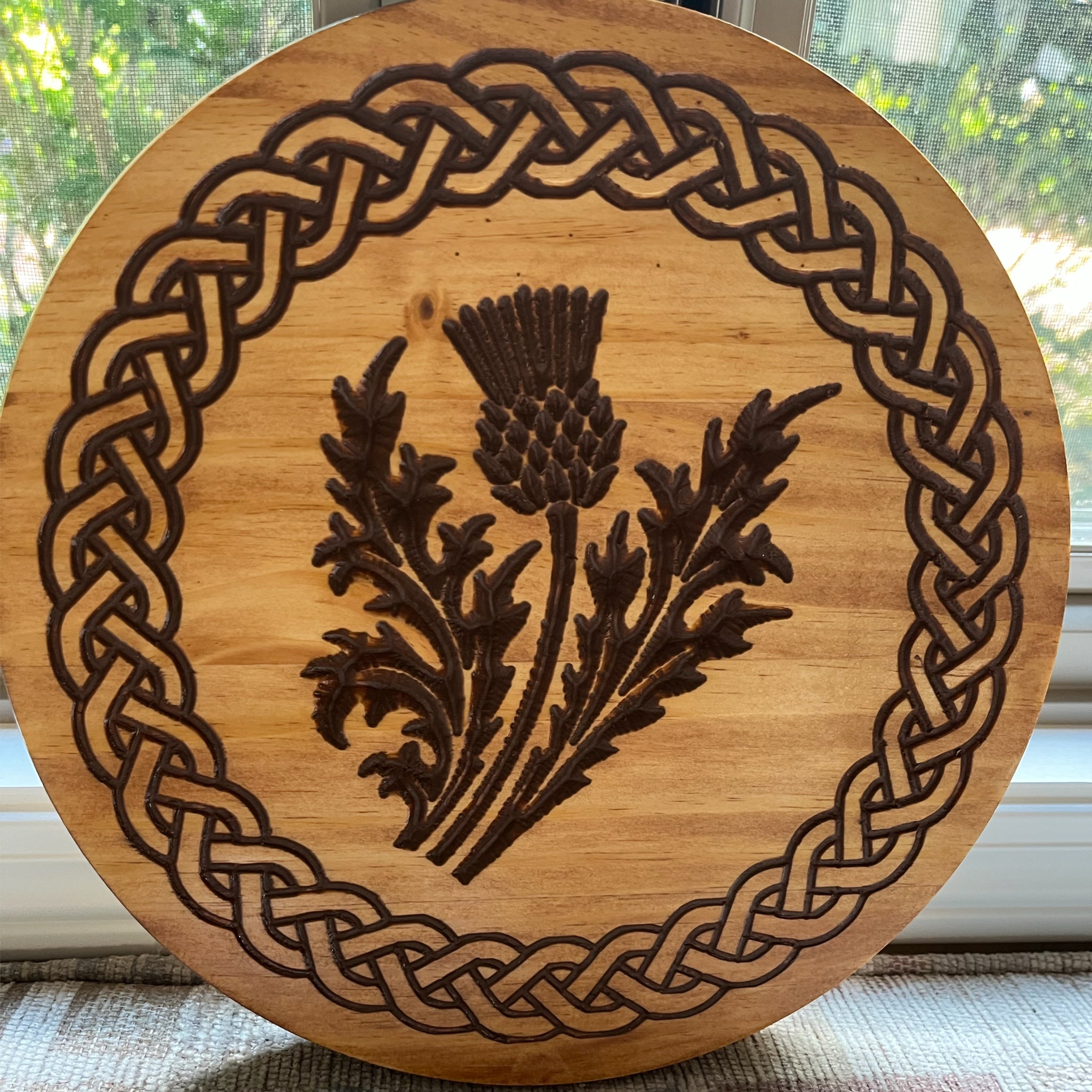 A round wooden plaque or coaster featuring a Celtic knot border and a central engraved thistle design, photographed in front of a window.