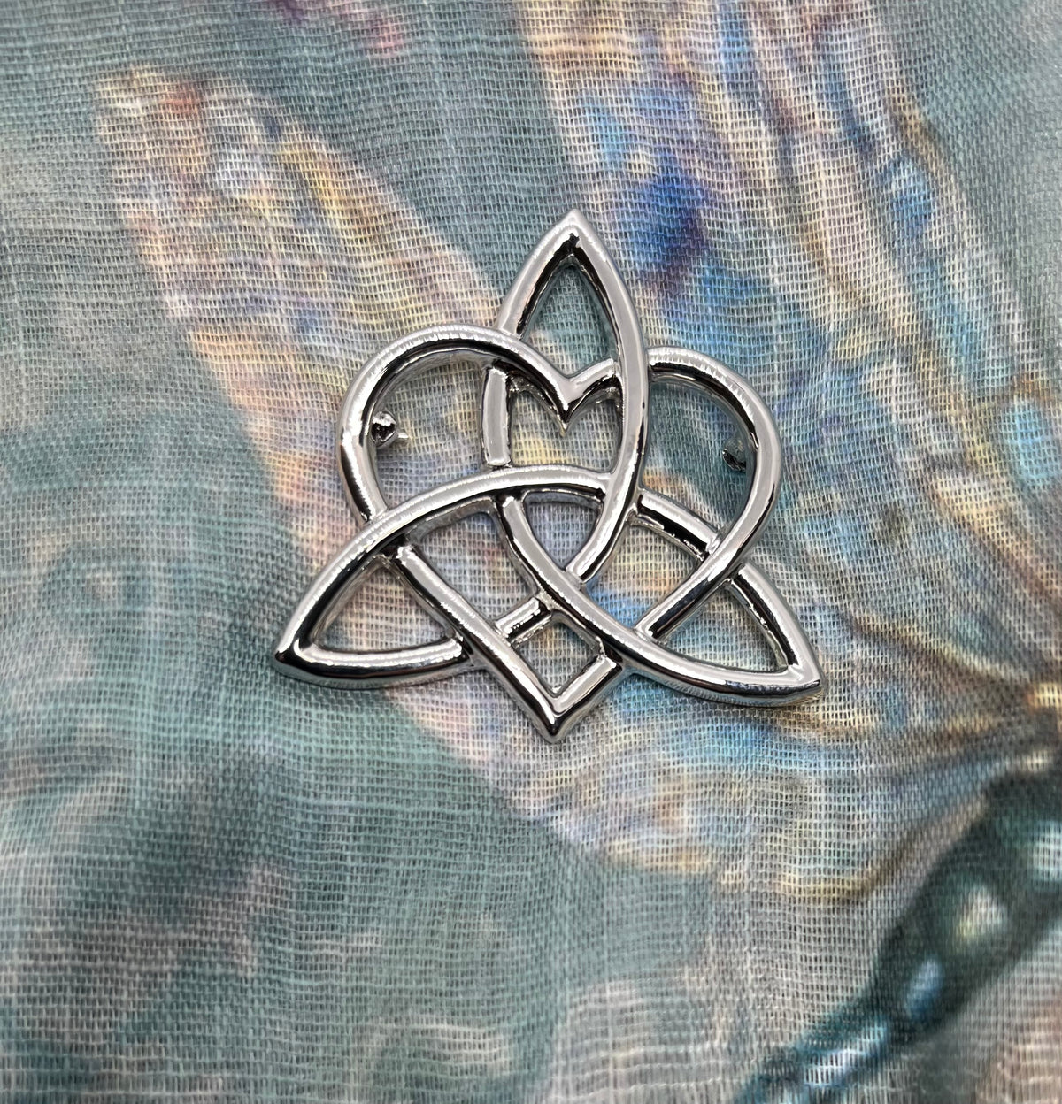 A silver-toned metal Celtic triquetra knot pin or brooch, displayed on a textured, blue-toned fabric background.