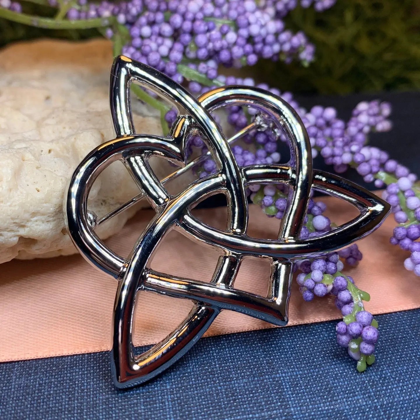 A polished pewter Celtic trinity knot brooch, styled on a peach and navy blue fabric surface with purple flowers and a stone in the background.