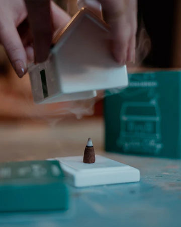 A close-up lifestyle shot of a person holding a white ceramic incense burner over a small incense cone on a white base, with teal product packaging in the background.