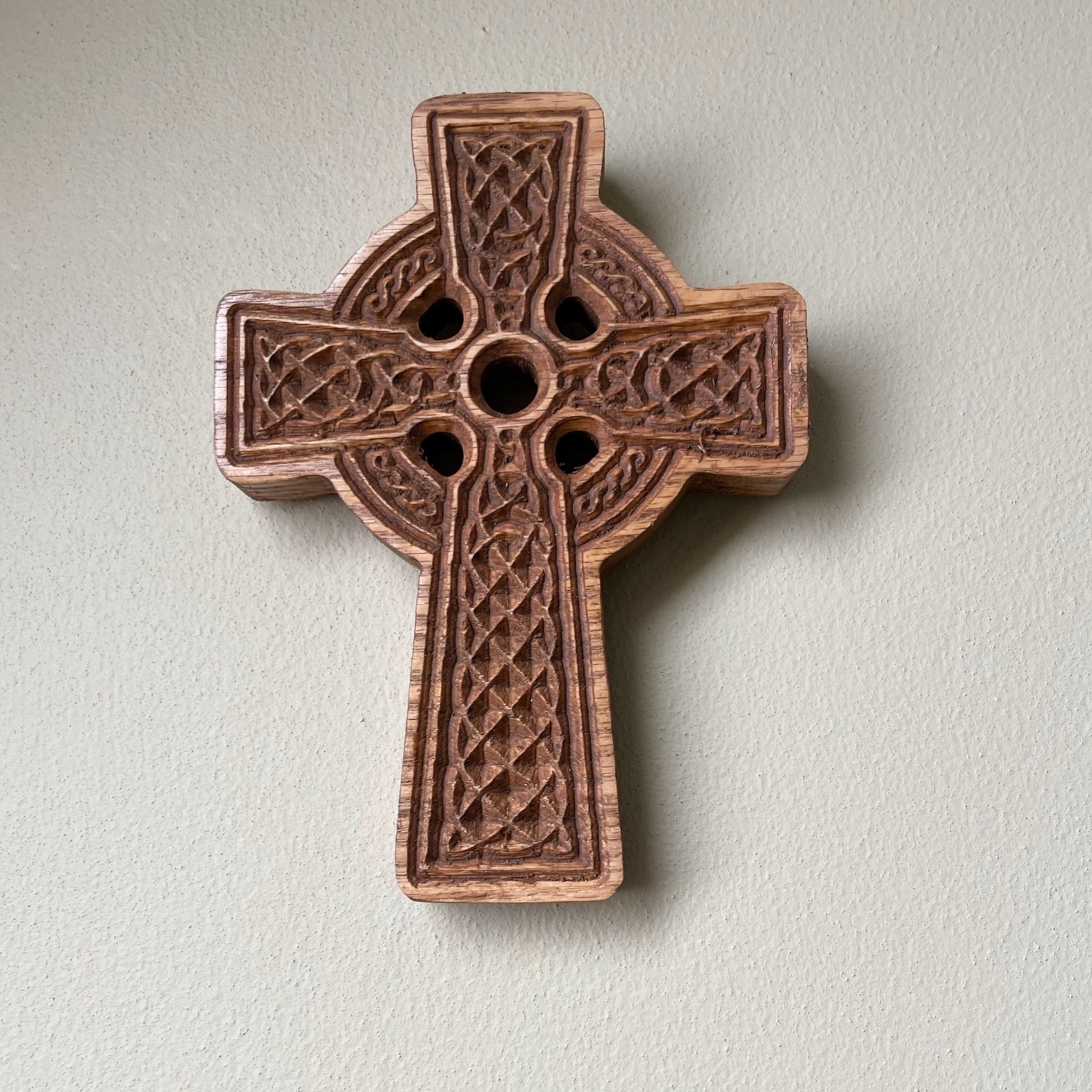 A carved wooden Celtic cross with intricate knotwork patterns and three circular cutouts, mounted on a textured off-white wall.