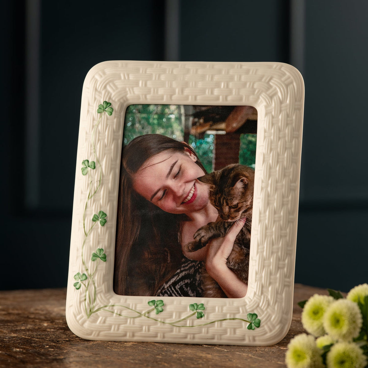 An off-white textured ceramic picture frame with green shamrock accents, displaying a photo of a woman and cat, set on a wooden surface with green flowers in the foreground.