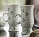 Two white ceramic mugs with woven pattern and clover design on a reflective surface.