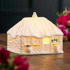 Cottage-shaped night light with a warm glow on a wooden surface with flowers.