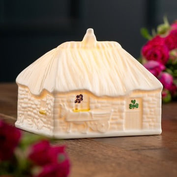 Cottage-shaped candle with a warm glow on a wooden surface with flowers in the background