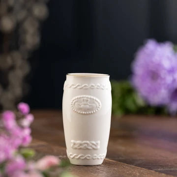 White Claddagh decorative vase on a wooden surface with blurred flowers in the background