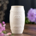 White decorative vase with intricate patterns on a wooden surface with blurred flowers in the background