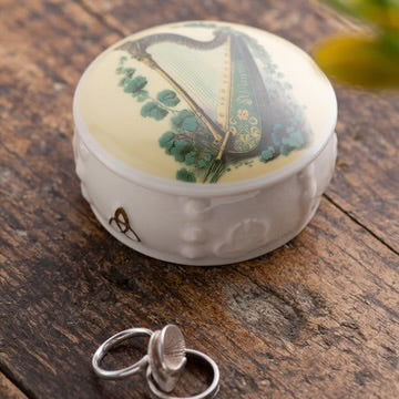 Round decorative box with a harp design on a wooden surface, accompanied by a silver ring.