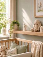 A styled nursery corner with a light wood crib, soft sage green pillows, a cream knit blanket, a plush bunny toy, a small shelf with a trailing green plant and a ceramic angel figurine, and a pale green wall with framed artwork.