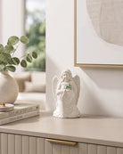 A small white angel figurine with wings and a green shamrock accent stands on a light beige console table in a styled interior setting.
