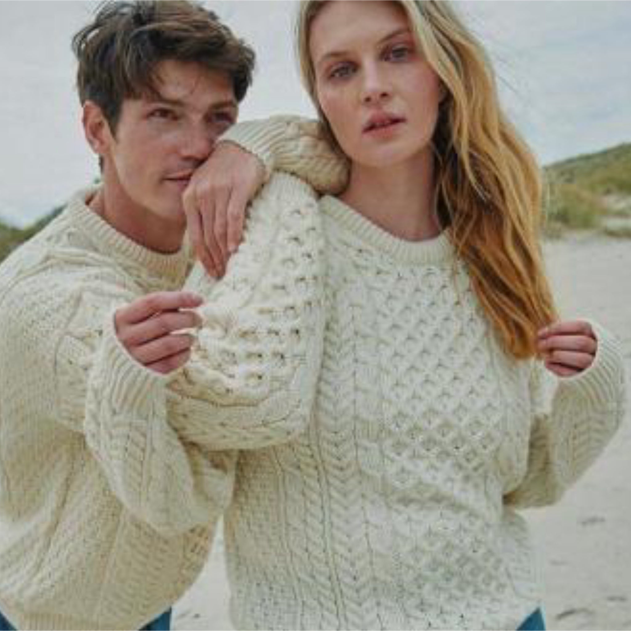 Two people wearing cream-colored Irish knitted sweaters on a beach.