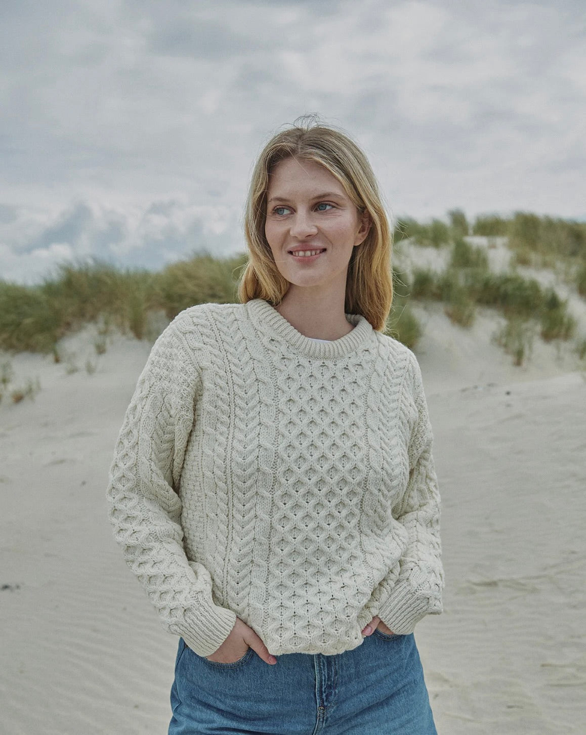 A woman with blonde hair stands on a sandy beach wearing a cream-colored cable-knit sweater and blue jeans, with dunes and a cloudy sky in the background.