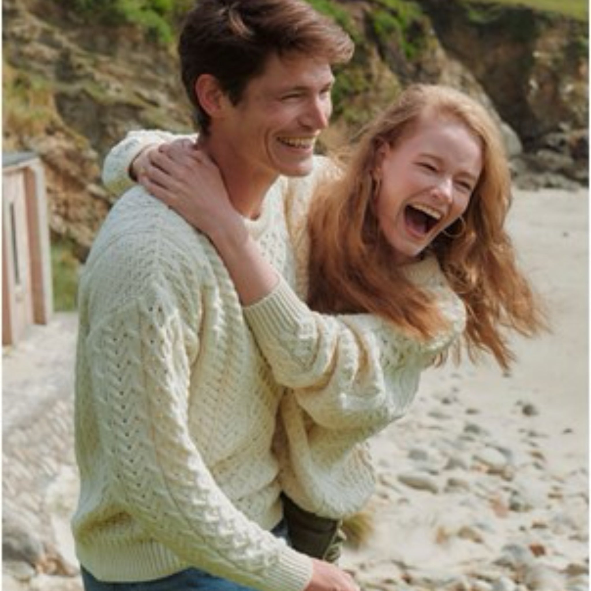 Two people in light-colored sweaters standing on a pebbly beach.