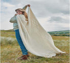 Woman holding a large white knitted blanket in a grassy field with a scenic background
