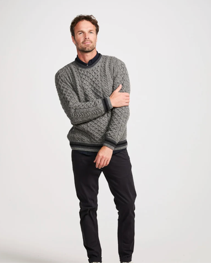 A man wearing a gray cable-knit crewneck sweater with black trim over a dark collared shirt and black trousers, standing against a light gray studio background.