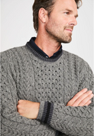 A man wearing a gray cable-knit sweater with a dark navy collar and matching striped ribbed cuffs, posed with his arms crossed against a light gray background.