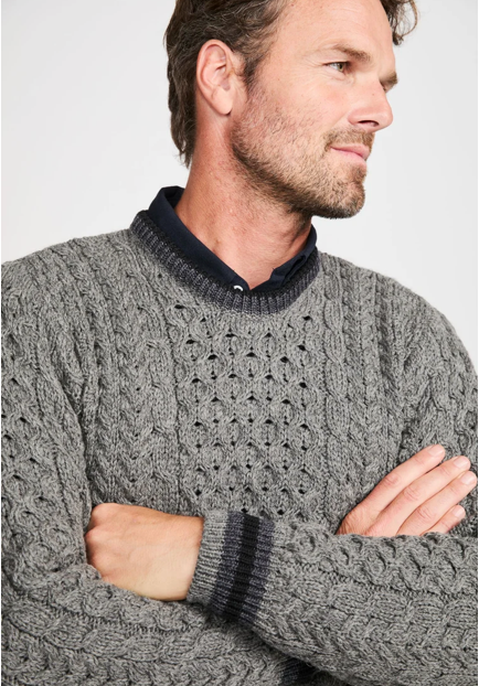 A man wearing a gray cable-knit sweater with a dark navy collar and matching striped ribbed cuffs, posed with his arms crossed against a light gray background.
