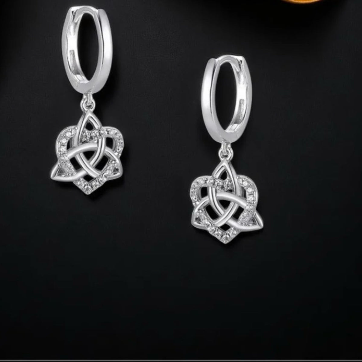 A pair of silver huggie hoop earrings with a dangling Celtic knot triquetra charm embellished with small clear stones, displayed on a dark black background.